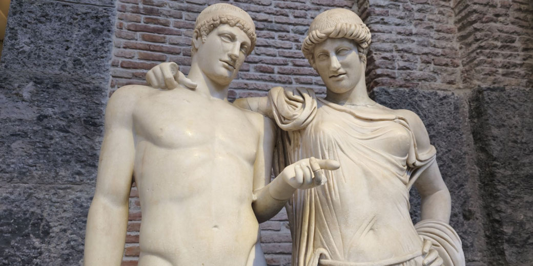 Marble statues of a nude male and a robed female figure standing closely, with the woman pointing and the man leaning on her shoulder, set against a stone and brick wall.