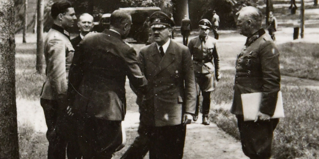 Black-and-white photo of uniformed men outdoors during WWII, with one man shaking hands in the center of the group.