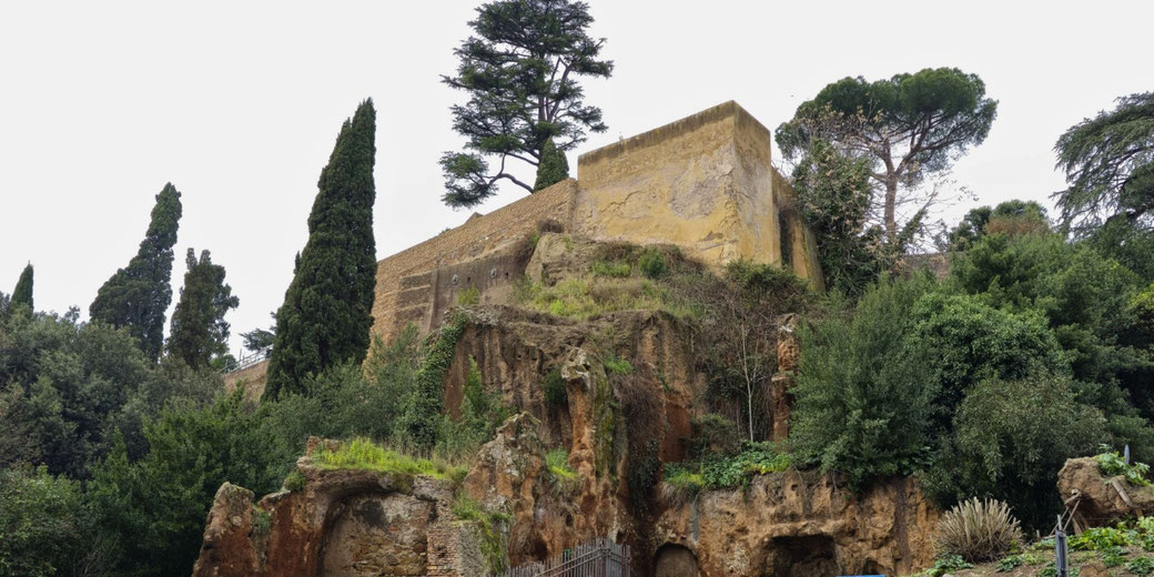 Ancient stone structure built into a rocky hillside, surrounded by lush greenery and tall cypress trees, with weathered walls and caves below.