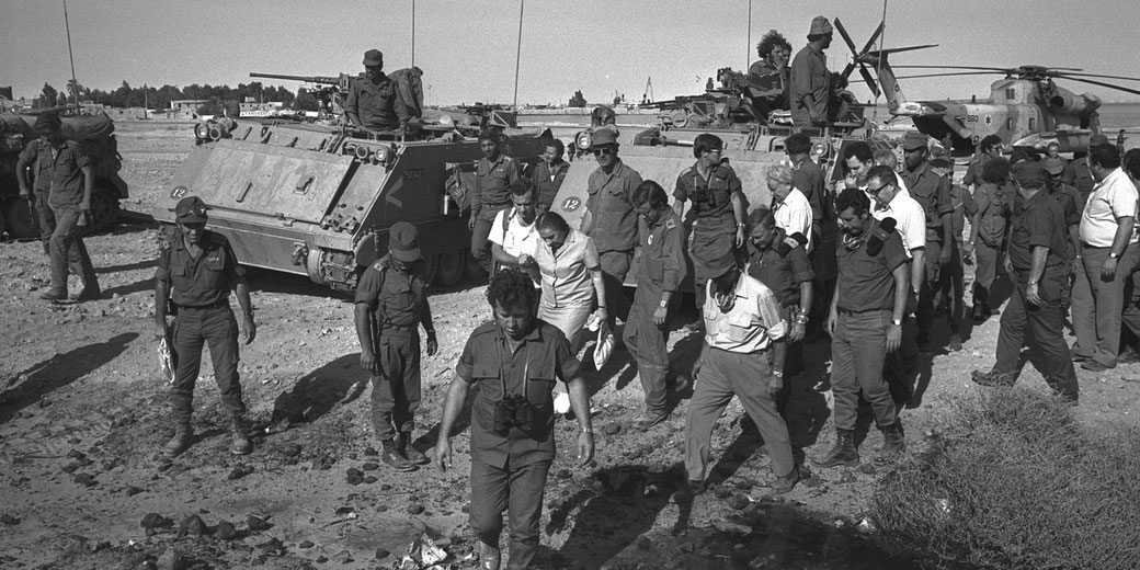 Military personnel and civilians gather around armored vehicles and helicopters in a desert setting, examining the ground.