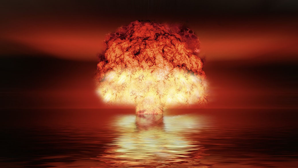 A fiery mushroom cloud rises over water, casting intense red and orange reflections across the surface.