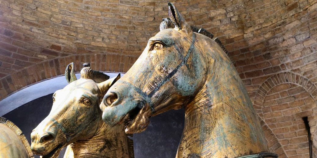 A close-up of two golden-bronze horse sculptures with a weathered patina, set against a brick archway.
