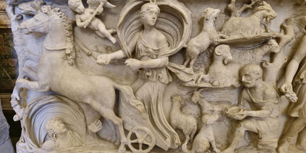 Ancient relief showing a woman in a chariot, led by horses, surrounded by animals, a child with wings, and a seated man offering a bowl, all carved in detailed marble.