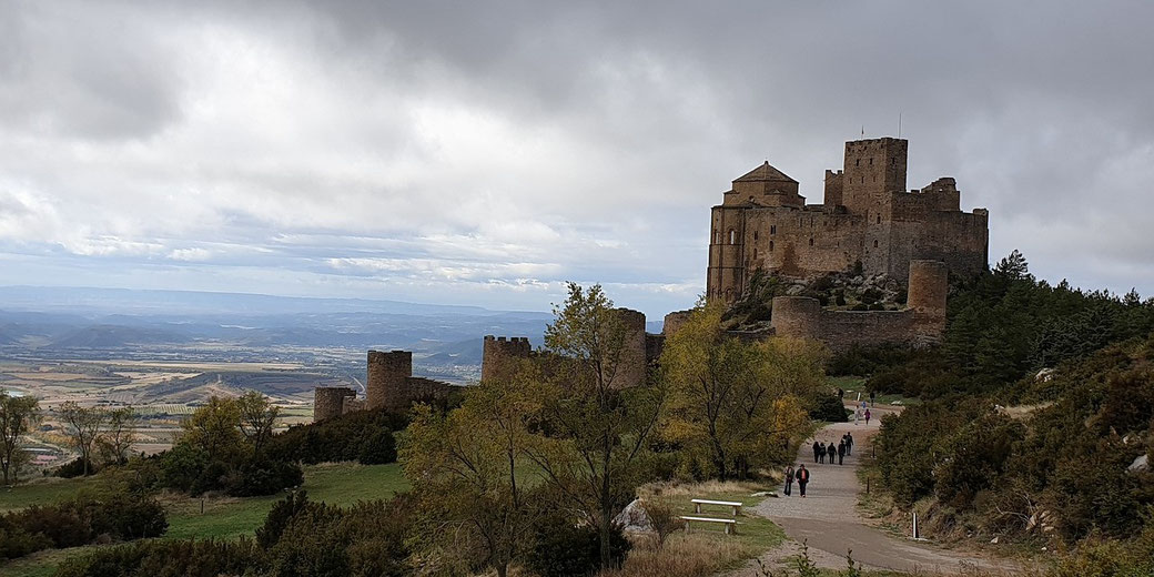 A medieval stone castle stands on a hilltop overlooking a vast valley, with visitors walking along a winding path below.