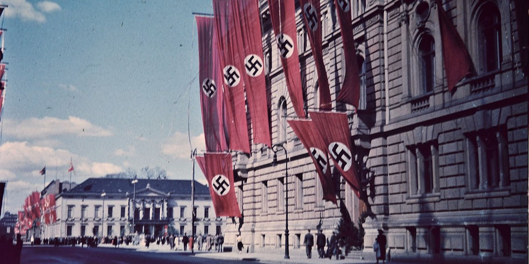 Large Nazi flags hang from a stone government building along a street with people walking and distant red banners visible.