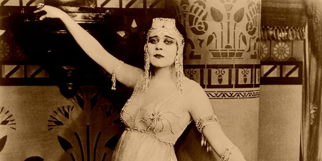 A vintage sepia portrait of a woman in decorative costume, wearing a beaded headdress and armbands, posing with one arm raised against patterned interior décor.