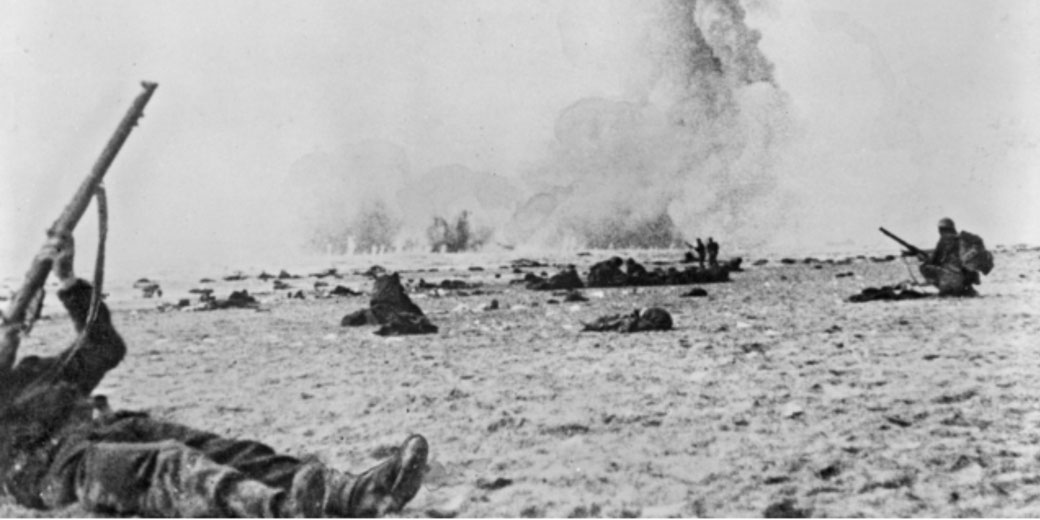 Soldiers take cover on a sandy beach as explosions erupt in the distance during an intense World War II battle.