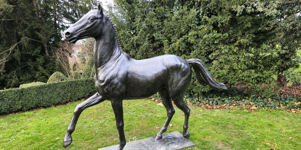 A bronze statue of a horse in a prancing pose, set in a lush green garden with trimmed hedges and tall trees in the background. The statue has a sleek, detailed, and polished finish.