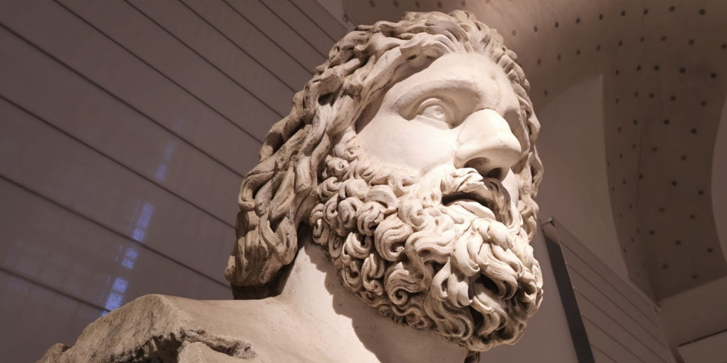 Low-angle view of a marble statue with flowing hair and a thick beard, gazing upward with a solemn, powerful expression.