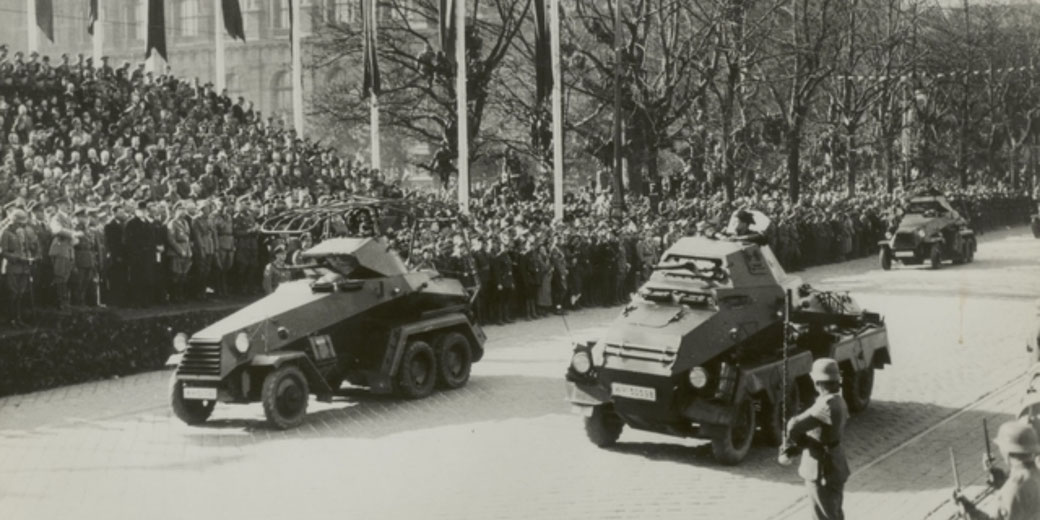 Armored military vehicles roll through a city street during a large parade, watched by tightly packed crowds and soldiers in uniform.