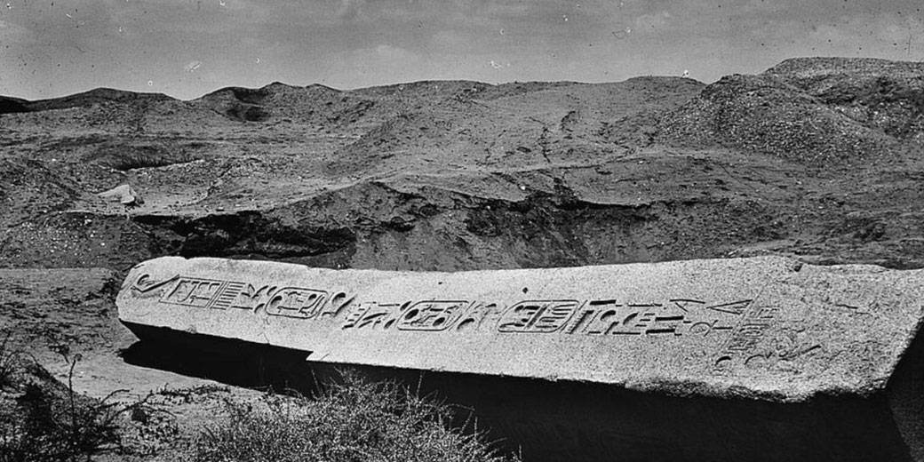 A fallen stone obelisk fragment lies in the sand, carved with clear ancient Egyptian hieroglyphs, surrounded by a barren desert landscape.