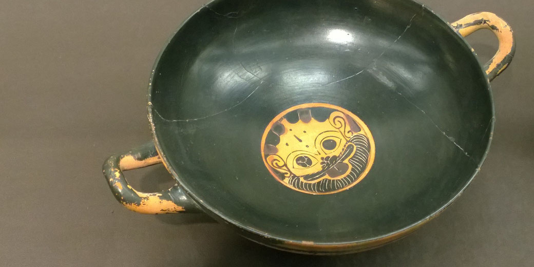 Ancient Greek black-glazed kylix with a painted gorgon face at the base, featuring two handles and visible wear and cracks.