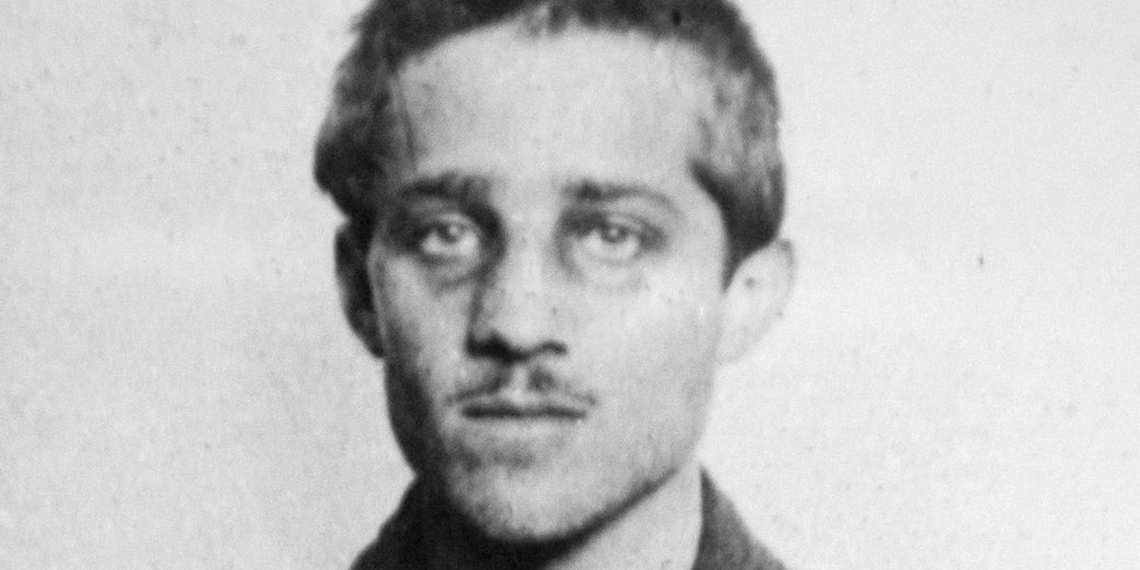 A black-and-white photograph of a young man with short hair and a mustache, looking directly at the camera with a serious expression.