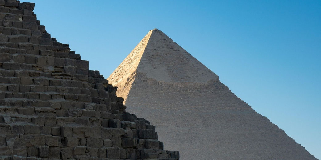 Ancient stone pyramids rise against a clear sky, with a partially ruined structure in the foreground and a large intact pyramid behind it in bright sunlight.