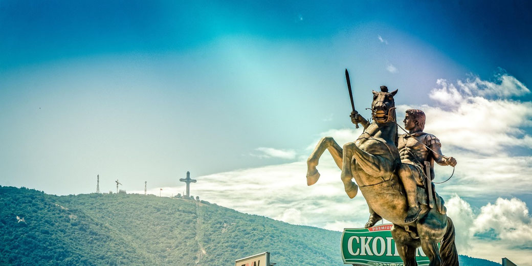 A bronze equestrian statue of a warrior holding a sword rises against a blue sky, with a mountain and cross in the background.