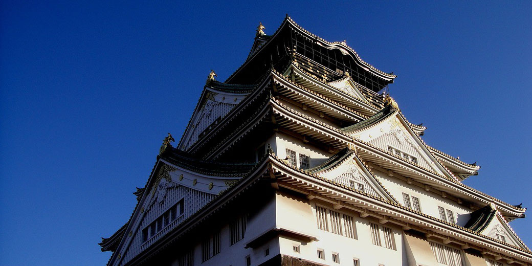 A multi-tiered Japanese castle with white walls and green-tiled roofs is shown from a low angle against a deep blue sky.