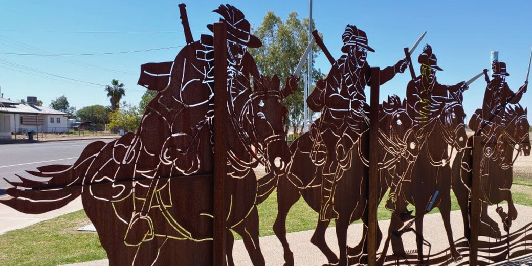 Metal silhouettes of mounted soldiers with raised swords form a striking roadside memorial, capturing a moment of cavalry charge against a bright blue sky and rural backdrop.