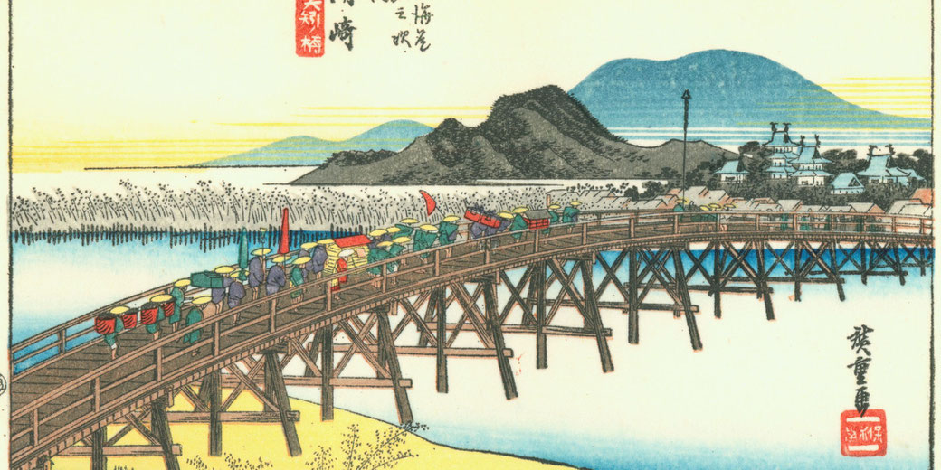 A traditional Japanese woodblock print shows people crossing a wooden bridge with mountains and a town in the background at sunset.