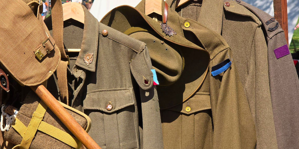 Close-up of World War I military uniforms and gear on hangers, including jackets, a hat, a satchel, and a canteen with colored unit patches.