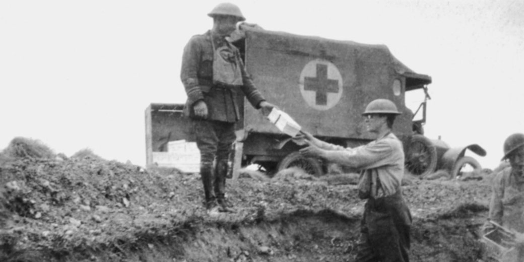 Black and white photo of a soldier handing supplies to another in a trench, with a World War I ambulance marked by a red cross in the background.