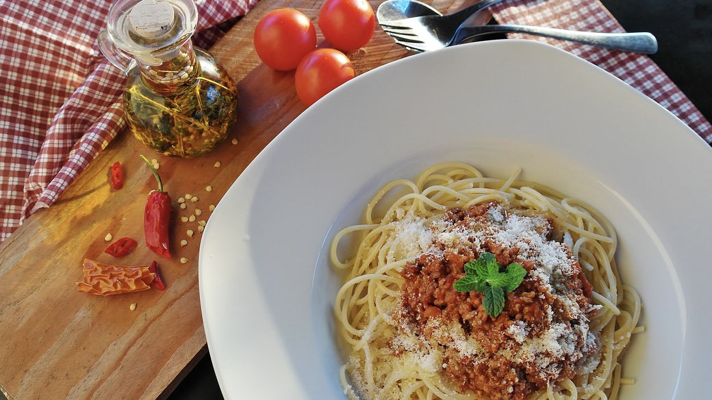 A plate of spaghetti with meat sauce, topped with grated cheese and a mint leaf. Surrounding ingredients include cherry tomatoes, chili peppers, and a bottle of infused oil on a wooden board with a checkered cloth.