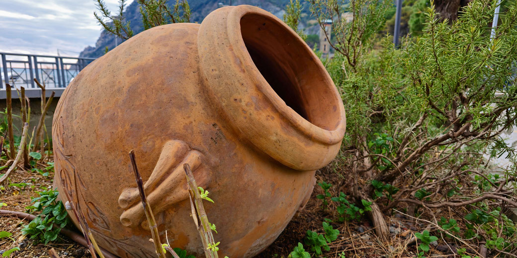 A large, weathered terracotta pot lies on its side in a garden surrounded by plants and shrubs, with a distant backdrop of hills, railings, and a cloudy sky.
