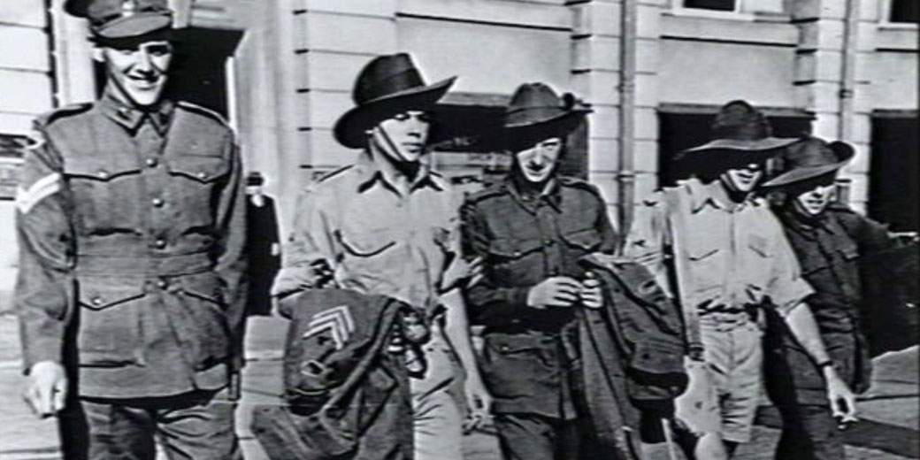Group of smiling soldiers in uniform and slouch hats walk together outside a building, holding jackets.