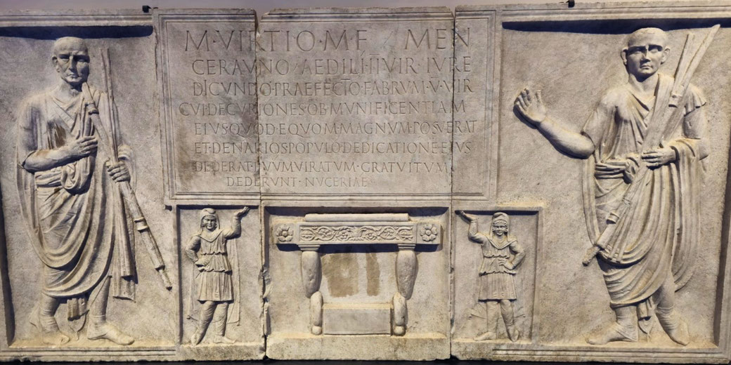 Ancient Roman relief showing two men in togas holding staffs, flanked by smaller figures, with a central Latin inscription and an altar below.