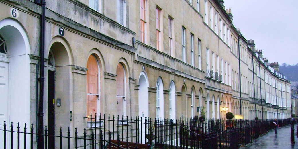 A row of elegant Georgian-style terraced houses with arched doorways and tall windows lines a wet, reflective street.