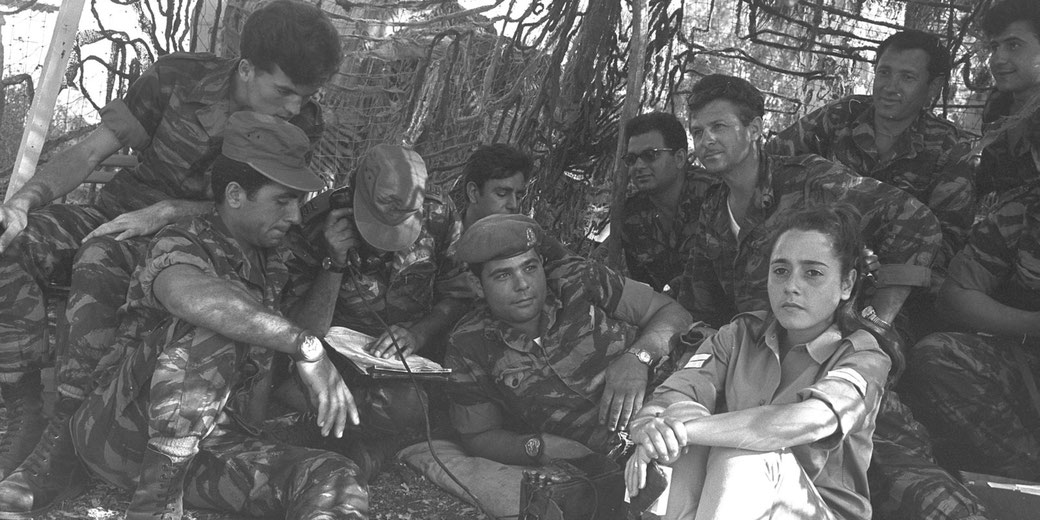 A group of soldiers in camouflage uniforms sits under netting, gathered around a radio and notes, with one woman seated at the front.