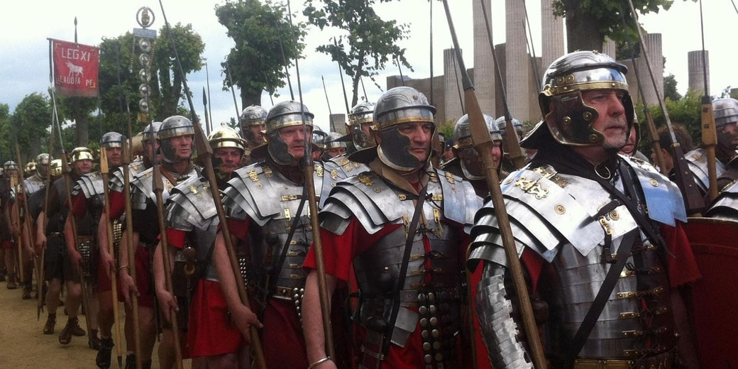Roman legion reenactors march in formation wearing metal armour and helmets, carrying spears and shields during a public historical event.