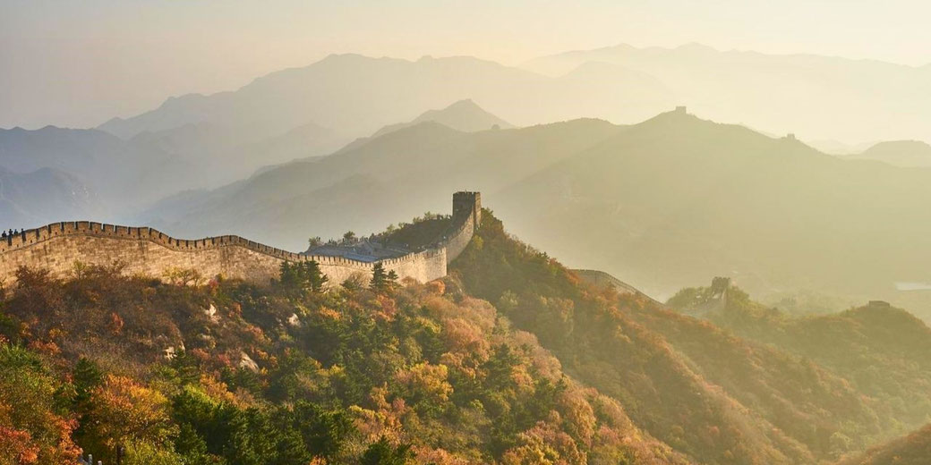 The Great Wall of China winding through misty mountains. The stone wall stretches into the distance, flanked by lush autumn foliage and watchtowers overlooking the rugged terrain.
