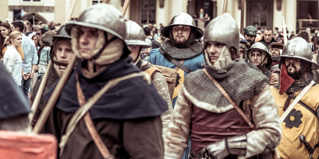 A group of people dressed as medieval soldiers march in a historical reenactment, wearing chainmail, helmets, and carrying weapons.