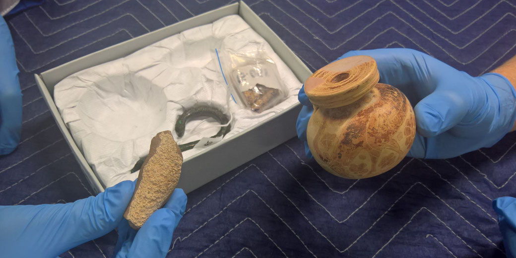 Gloved hands hold ancient artifacts: a small patterned pottery vessel and a fragment of another item. Nearby, a box contains other relics, including a metal piece and wrapped items, on a protective blue surface.