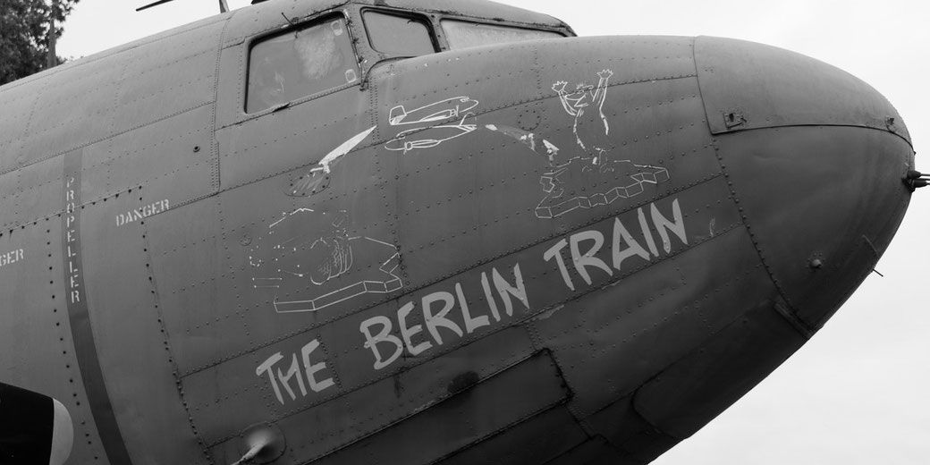 Black-and-white image of a military aircraft nose with "The Berlin Train" painted alongside cartoon art of a bear and parachuting supplies.