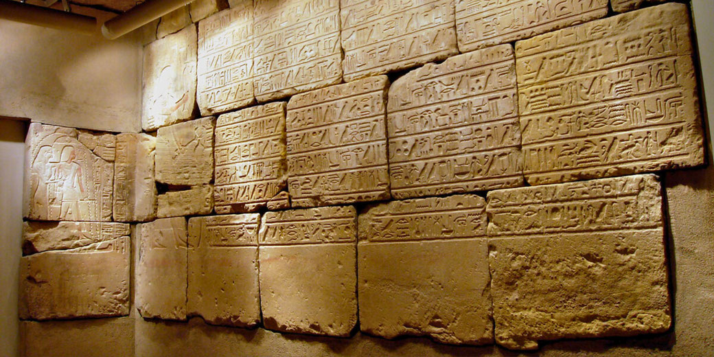 Ancient Egyptian wall with rows of hieroglyphics carved into large stone blocks, lit by soft indoor lighting. A faint figure is seen on the left side.
