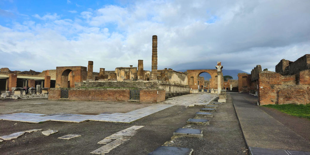 The Forum of Pompeii, surrounded by ancient ruins, columns, and arches, with Mount Vesuvius in the background under a partly cloudy sky. The site served as the city's civic and social hub.