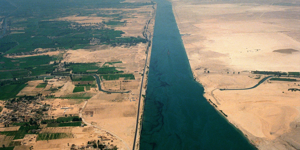 A long, straight waterway divides lush farmland on one side and arid desert on the other, viewed from above.