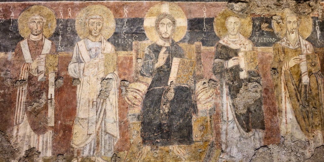 Ancient fresco showing five haloed religious figures, likely early Christian saints and Christ, each holding books, with worn and aged surface details.
