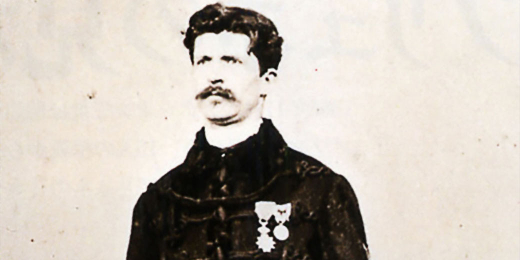 A vintage photograph of a man with a mustache in dark formal clothing displays two medals pinned to his chest against a plain background.