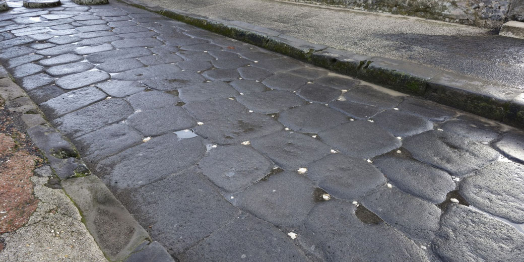 Close-up of an ancient Roman stone road with worn, uneven basalt paving stones, slightly wet from recent rain.