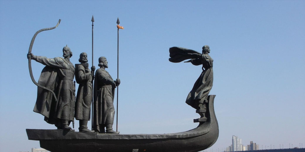 Four bronze figures stand on a stylized boat, one drawing a bow and another with arms outstretched, set against a riverside cityscape.