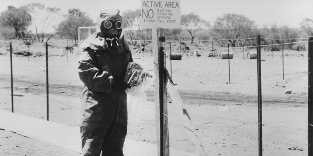 A person in full protective gear and gas mask stands near a warning sign in a fenced desert test area labeled “ACTIVE AREA NO EATING DRINKING SMOKING.”