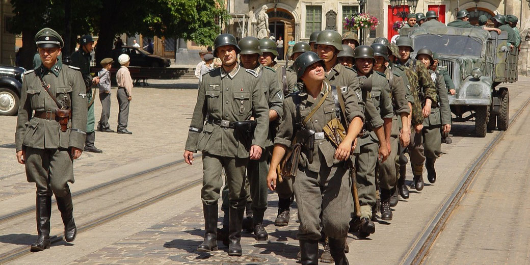 Group of men in WWII-era German military uniforms march down a cobblestone street during a historical reenactment.