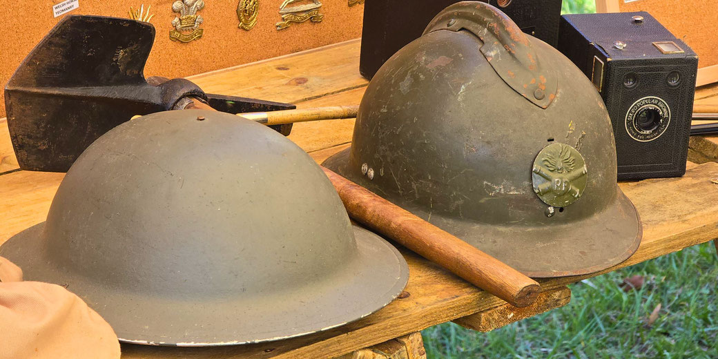 Two WWI-era Allied helmets, a British Brodie helmet and a French Adrian helmet, on a wooden table with a shovel, vintage camera, and military badges in the background.
