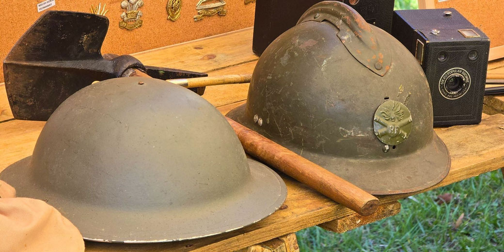 Two WWI-era Allied helmets, a British Brodie helmet and a French Adrian helmet, on a wooden table with a shovel, vintage camera, and military badges in the background.
