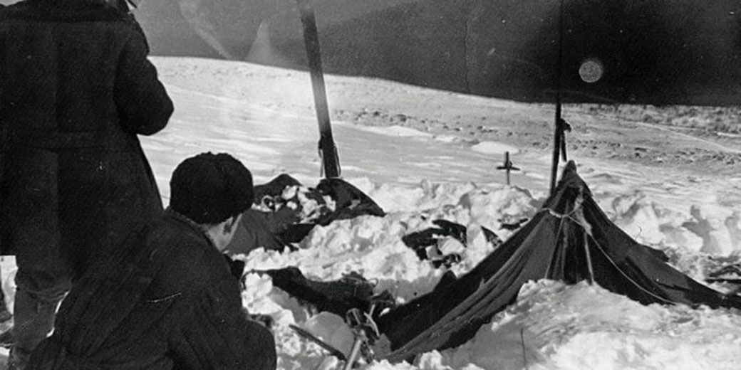 Black-and-white photo shows people examining a snow-covered tent in a remote, snowy landscape with scattered gear and ski poles.