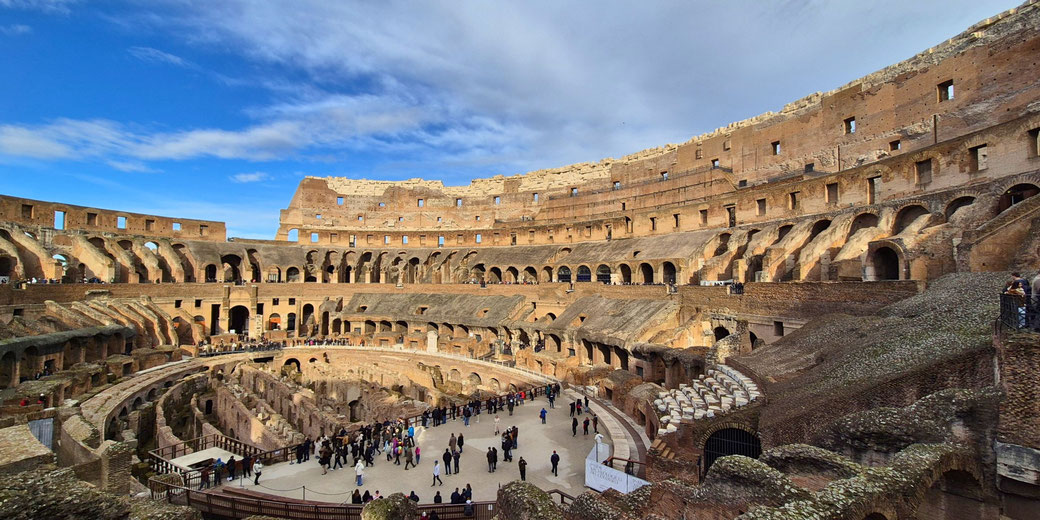 the interior of the Colosseum in Rome, showcasing its ancient ruins, hypogeum, and tiered seating. Tourists explore the vast amphitheater, once used for gladiatorial combat.