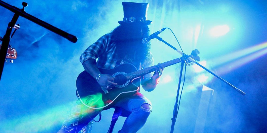 A musician in a top hat and flannel shirt plays acoustic guitar on stage under dramatic blue and purple lighting.