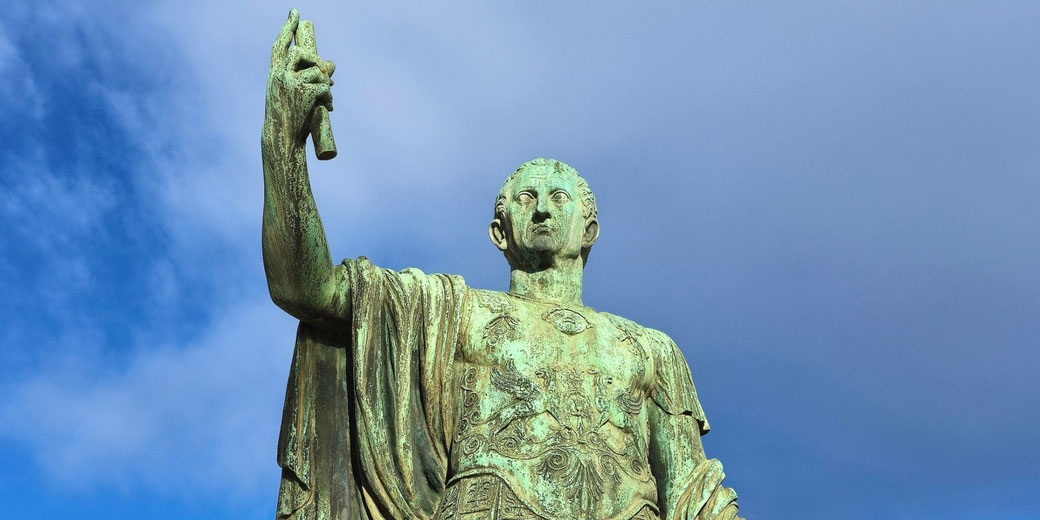 A green patina bronze statue of a Roman figure in armor raises a scroll, seen from a low angle against a cloudy sky.